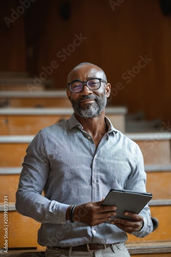 A professional Black male teacher in his 50s with dark skin and glasses holds a tablet in a college lecture hall auditorium.