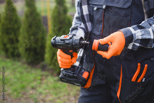person holding power drill outdoors wearing orange gloves and black jacket for home improvement project in garden setting close-up practical and serene mood natural light clean background.