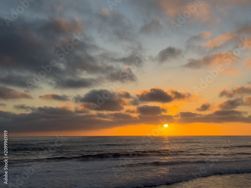 Obraz na plátně Orange and golden hues fill the evening sky and clouds during a dramatic sunset over the Pacific Ocean in Carlsbad, California