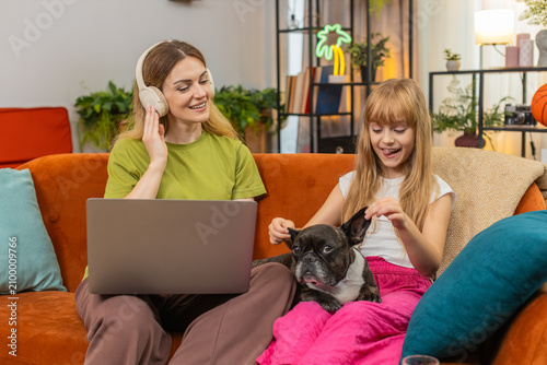фотографія Mother and daughter with French bulldog at home use wireless headphones play lo fi music while laptop work