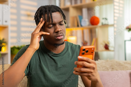African American young man on home sofa looking at smartphone with sad face after bad news and online game loss. Black guy on home sofa feels upset while reading screen and reacting to online defeat.