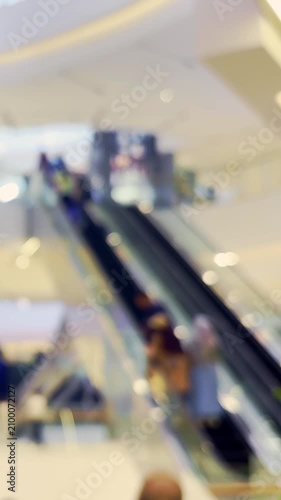 Soft focus view of shoppers and escalators inside a bright modern retail shopping mall.