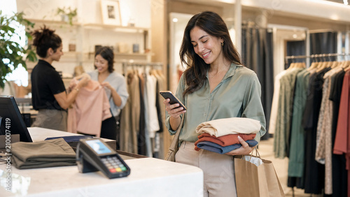 Woman shops in a clothing store, holding folded garments and using smartphone near checkout, showing retail fashion and mobile payment.