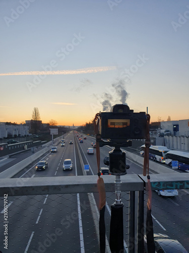 A camera mounted on a tripod frames a multi-lane highway at sunset, with cars and buses moving toward the city horizon, conveying travel, commute, motion and peaceful dusk light.