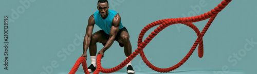 Muscular Black male athlete slamming red battle ropes creating rippling waves during training. High intensity fitness workout on teal background. Panoramic banner with copy space for text