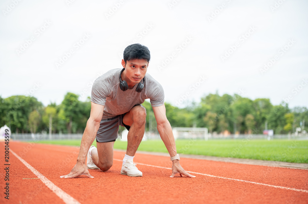 Fototapeta premium Asian man sprinter focused while crouching in start position ready to run on track in sports stadium