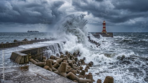 Massive ocean storm waves crashing over a concrete breakwater pier and a red lighthouse under dramatic dark storm clouds