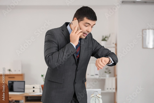 Stressed businessman with wristwatch talking by mobile phone in office. Time ...