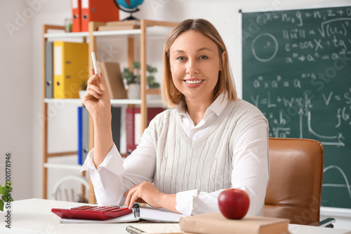 Mature female math teacher with chalk sitting at table with calculator in cla...