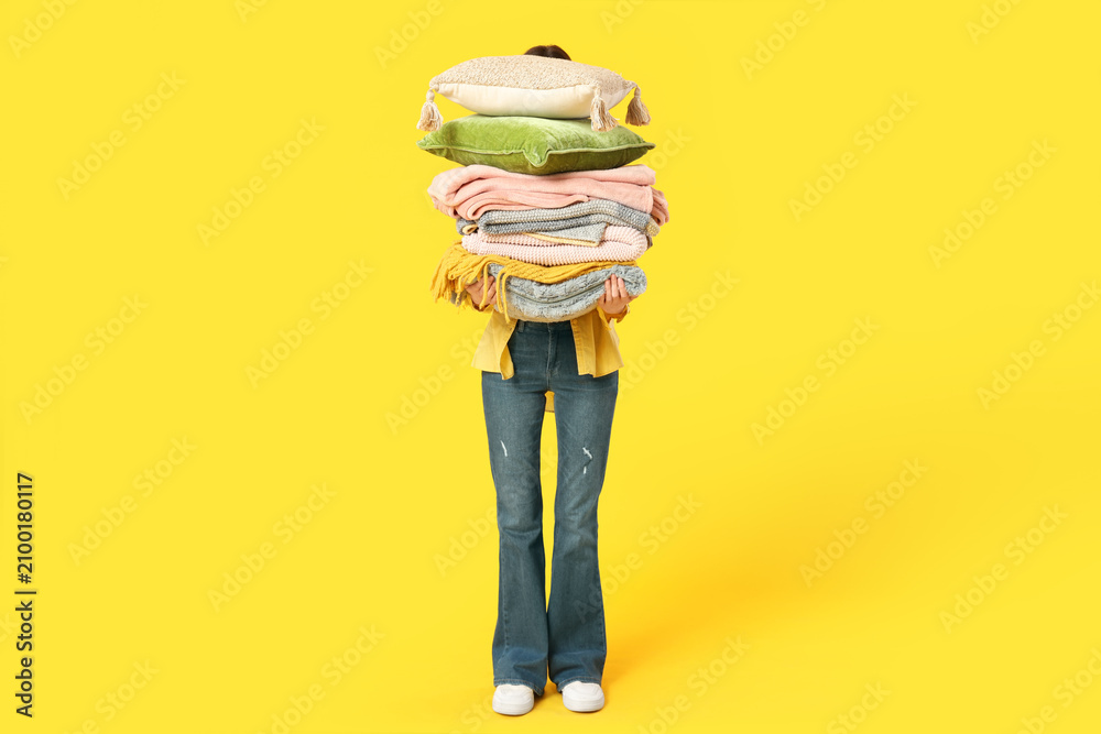 Naklejka premium Beautiful young woman with stack of different blankets and pillows on yellow background