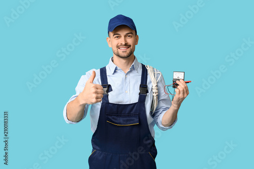 Portrait of male electrician with multimeter and wires showing thumb-up on bl...