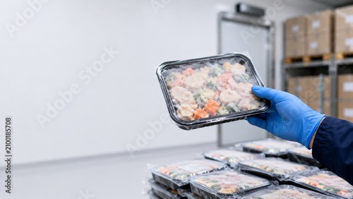 Hand with blue glove holding processed food tray in warehouse checking frozen meal product at food factory, ensuring quality and safety in industrial storage environment