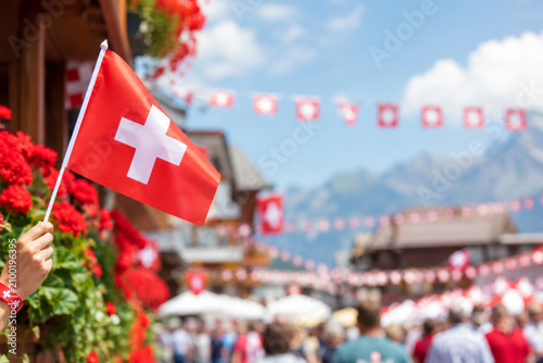 Hand holding a Swiss flag during celebration with alpine village and decorations in the background for Swiss National Day.