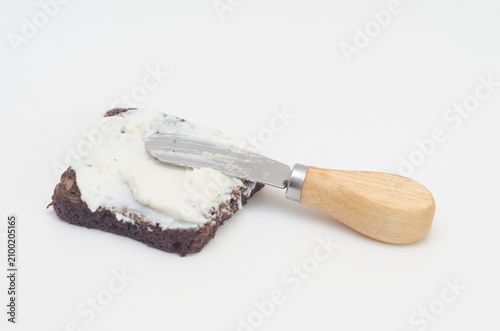Ricotta cheese spread on a slice of brown bread and butter knife on white background.