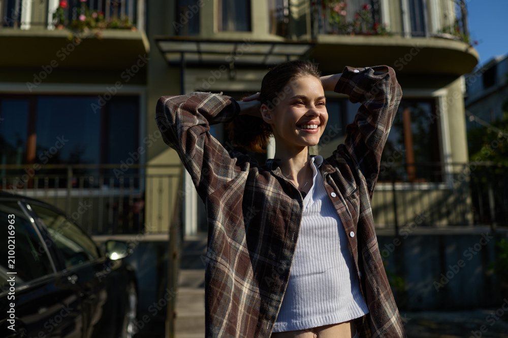 Fototapeta premium Casual outdoor portrait of a smiling woman wearing a plaid shirt, enjoying daytime sunlight, relaxed pose in front of a residential building, lifestyle mood