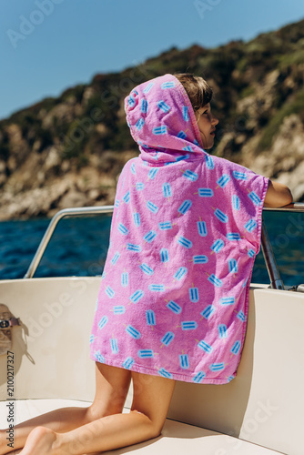 Child in a pink polka dot towel poncho sits on a boat deck looking out at the sea. Summer sea holidays with kids and cosy after swim moments for family travel marketing