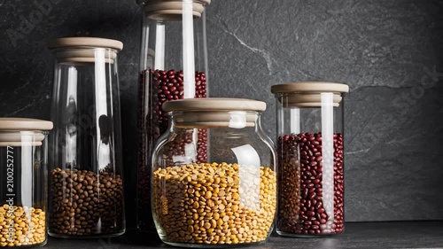 Assorted tall and rounded canisters filled with yellow split peas, brown pulses and red beans stand before a charcoal stone wall, creating dramatic modern pantry styling with generous copy space