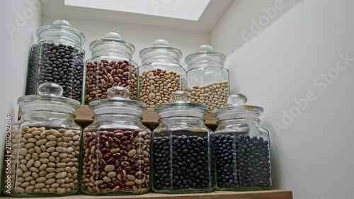 Two tier arrangement of lidded glass canisters containing black, cream and speckled beans rises beneath a bright skylight, giving a compact pantry corner a clean vertical structure and airy freshness