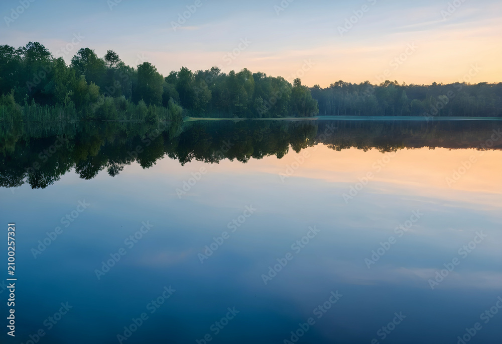 Obraz premium Calm lake with forest reflection at sunrise, tranquil nature landscape with clear sky and still water, the clear sky transitions from blue to orange and yellow hues