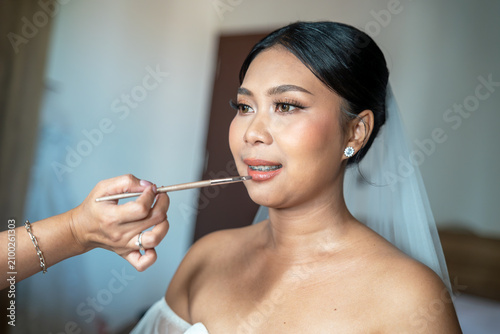 Foto Close up of an elegant Asian bride with braces getting her lipstick applied by a makeup artist during wedding day preparation