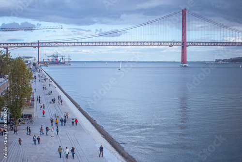 Quadro su tela People walking on the Tagus promenade near 25 de Abril Bridge in Lisbon, Portuga