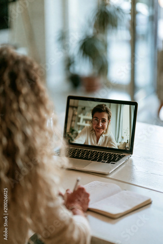 A candidate is sitting in a clean, minimalist home office, looking at a laptop screen where a recruiter's face is visible in a video call.