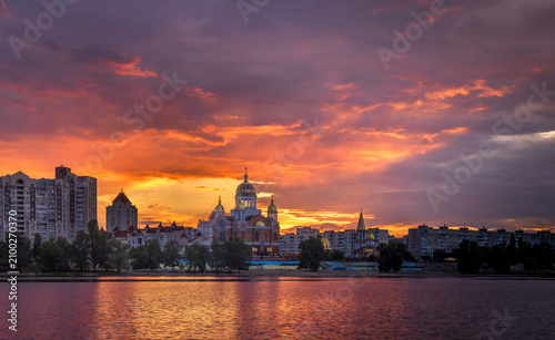 Vivid afterglow over Obolon Embankment in Kyiv, Ukraine. Dramatic purple and orange clouds illuminate the skyline and church, reflecting across the calm waters of the Dnipro River.