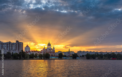 Golden sunset over Obolon Embankment in Kyiv. Rays of sunlight burst behind a church and city skyline, reflecting on the Dnipro River beneath dramatic storm clouds.