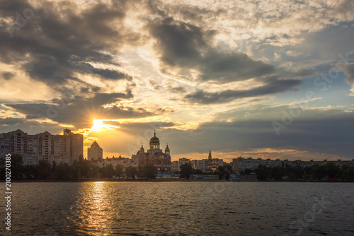 Golden sunset over Obolon Embankment in Kyiv. Rays of sunlight burst behind a church and city skyline, reflecting on the Dnipro River beneath dramatic storm clouds.