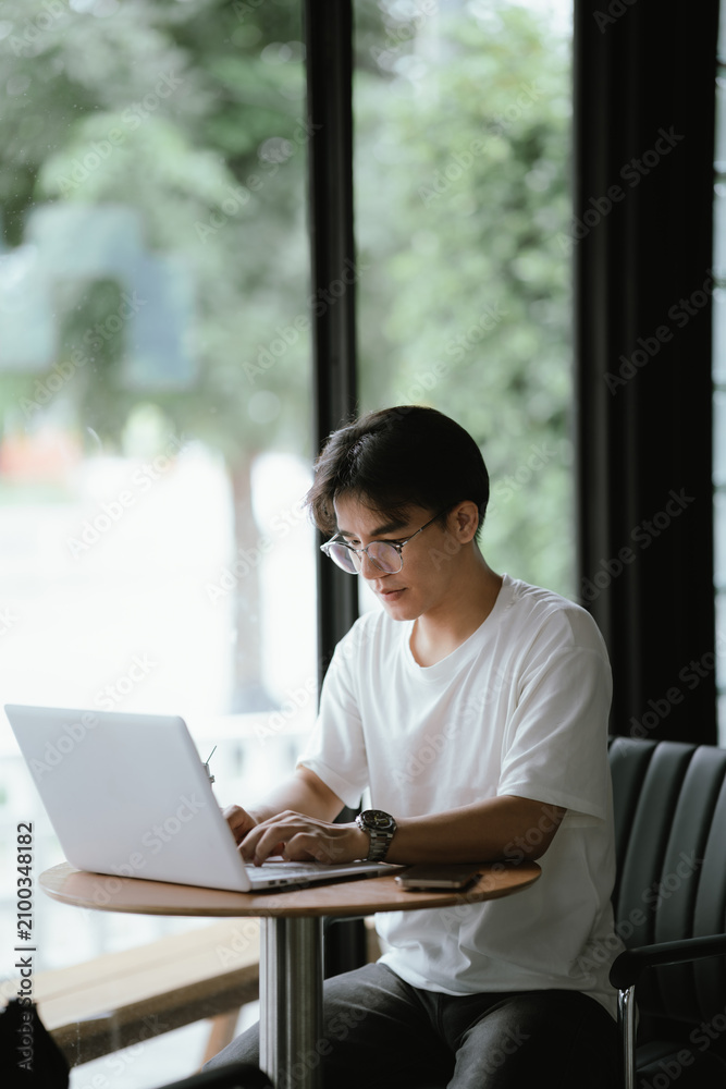 Fototapeta premium Young professional talking on smartphone while working on a laptop in a modern café, holding a coffee cup with natural window light.