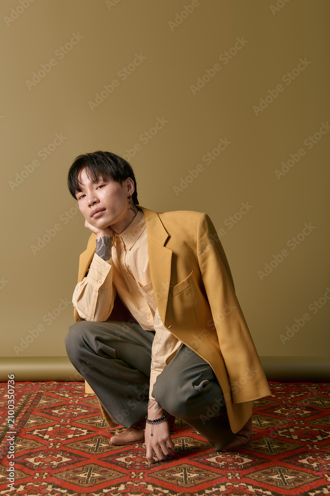 Fototapeta premium A handsome young Asian man squats thoughtfully in chic clothing, showcasing modern style and flair.