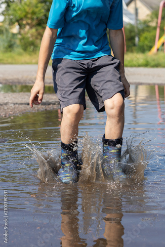 cropped image of a child in rubber boots joyfully jumping and splashing in a puddle after a summer rain. The freedom of childhood happily spending summer days, changeable weather, outdoor play.