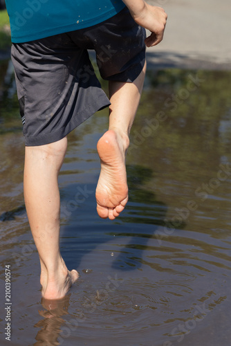 The sunlit bare feet of a boy playing in a puddle after a summer rain. Childhood freedom, sensory experience, exploring the world. Changeable weather. Outdoor play.