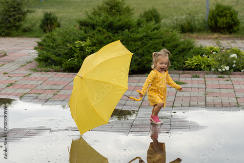 sweet little girl in rubber sandals, yellow dress, holding large umbrella, happily runs through puddle after summer rain. freedom of childhood, joyful emotions, exploring the world with curiosity.
