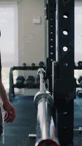 Vertical video: Entering man in gym wear standing by rack in small gym, preparing lift with barbell