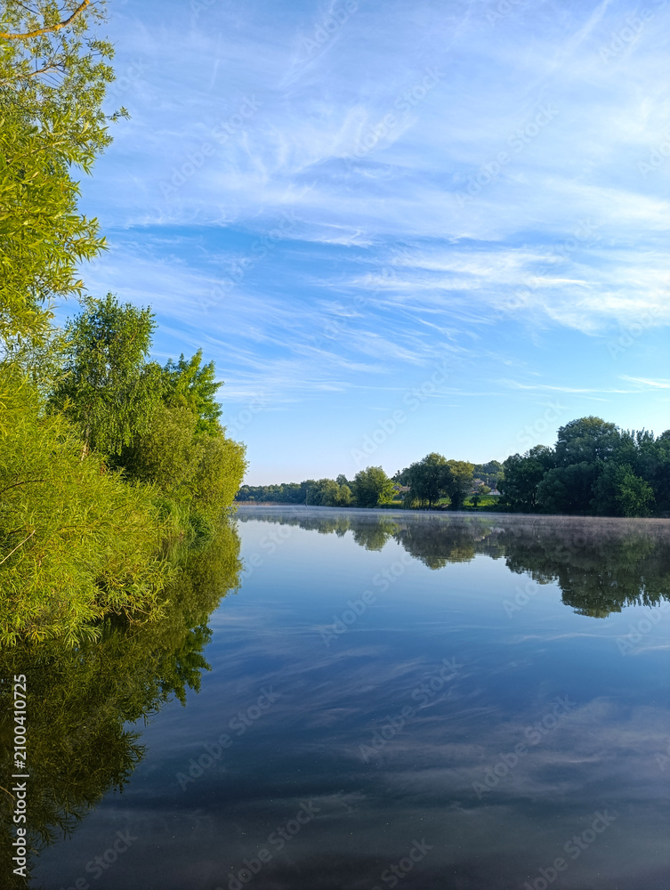Obraz premium Beautiful reflection of blue sky and wispy clouds in a calm river surrounded by green trees