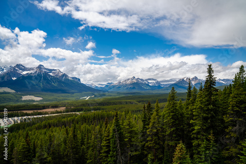 Panoramic view of snow-capped mountains rising above dense evergreen forest beneath dramatic summer skies in the Canadian Rockies, Alberta, Canada.