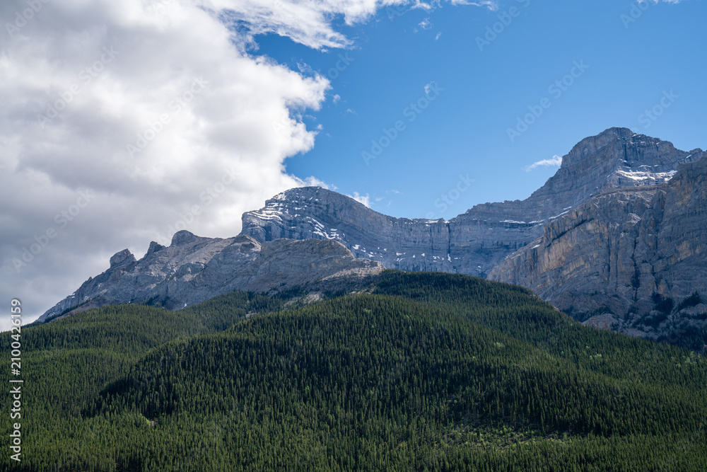 Fototapeta premium Layered limestone cliffs rise above dense evergreen forest under dramatic skies at Lake Minnewanka in Banff National Park, Alberta, Canada.