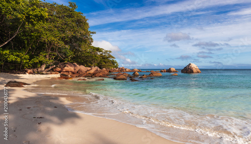 Anse Lazio, Praslin, Seychelles. Vibrant tropical beach featuring calm azure waters, large rock formations, and trees providing shade under a sunny blue sky
