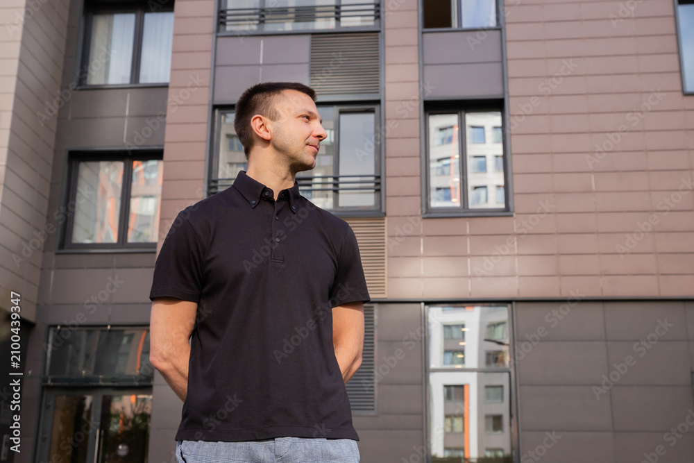 Obraz premium Caucasian man standing outdoors in front of a modern residential building. Young guy in a black polo shirt looking away. Urban lifestyle, professional portrait and city living concept.