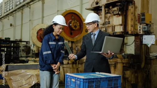 Asian manager and female worker in hard hats discuss and criticizing quality control of a machined metal part using a laptop. They stand near heavy machinery in a large industrial manufacturing plant 
