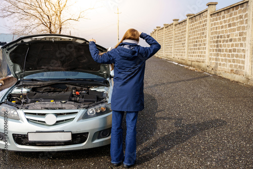 A stressed woman stands on the roadside with her head in her hands next to her broken-down car with an open hood, waiting for emergency road assistance.