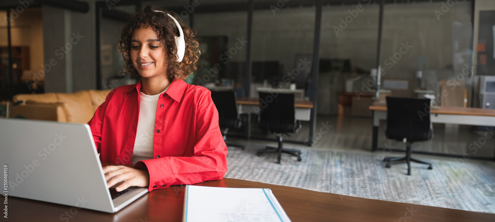 Obraz premium Young indian woman with curly hair working on laptop computer at desk in modern office or home workspace, wearing shirt and smiling. Latin or muslim arab student girl studying. Web banner, copy space