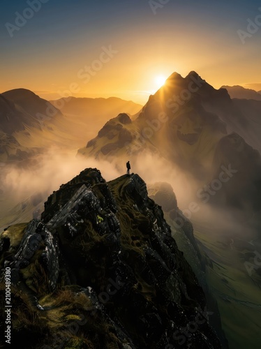 A dramatic vertical shot of a lone hiker standing at the absolute pinnacle of a rocky peak, celebrating a successful climb as the morning sun bursts over rugged mountain ridges.