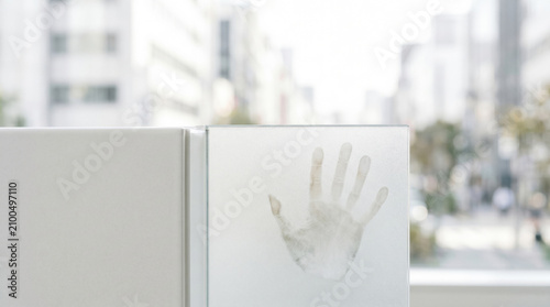 A single palm print appears on a glass partition near a window, suggesting touch and presence against a blurred city backdrop. Clean modern interior with soft natural light and negative space.
