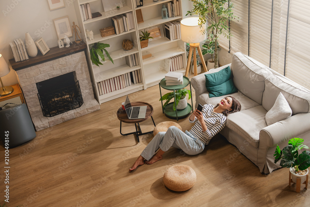 Obraz premium Overhead view of woman relaxing on floor with smartphone in cozy living room, fireplace and bookshelf background, laptop on table, warm home interior lifestyle scene with copy space