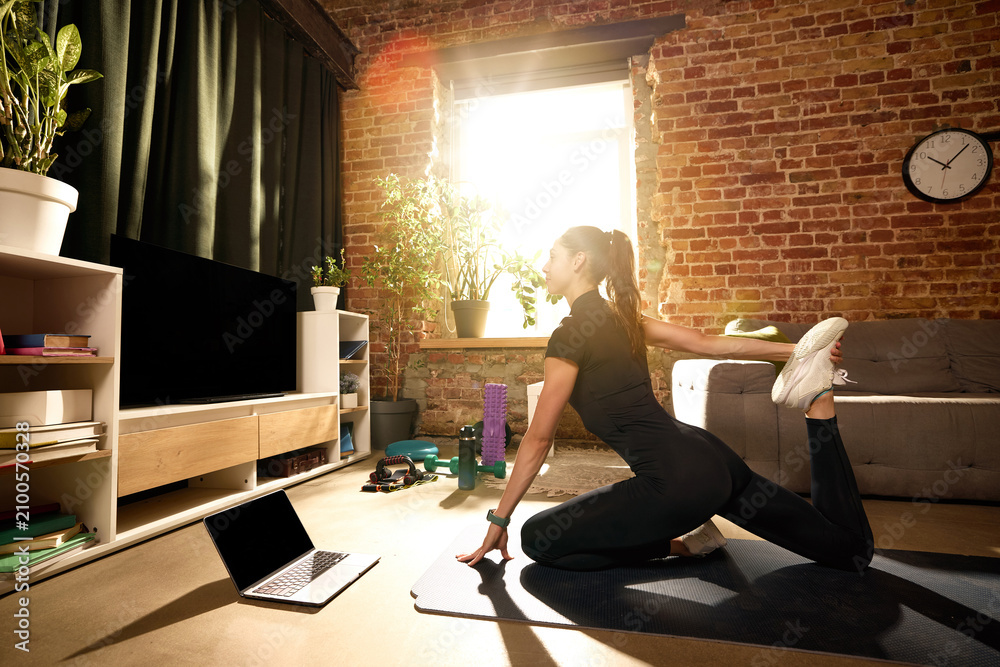 Obraz premium Woman stretching quadriceps on mat during online home workout. Concept of post-workout recovery, muscle flexibility, injury prevention, mobility improvement and virtual fitness coaching sessions.