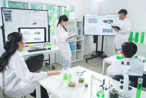 Laboratory scientist research team chemistry experiment presentation data chart microscope, group reviews report with glassware, green liquid, white coat, focused collaboration in bright lab