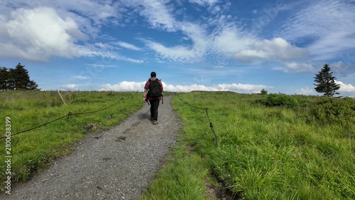 Hiker walking across Round Bald at Roan Mountain near North Carolina and Tennessee border, surrounded by open grassy ridges, distant clouds, and panoramic Appalachian mountain views