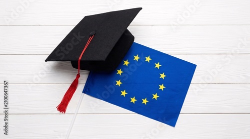 Black graduation cap with a red tassel resting beside a European Union flag on a textured white wooden surface symbolizing academic success